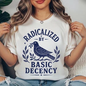 Op de afbeelding: Een wit T-shirt met korte mouwen en een donkerblauwe grafische print. Het ontwerp toont een vogel met een takje, omringd door bladeren, met de tekst RADICALIZED BY BASIC DECENCY in een opvallend, verweerd donkerblauw lettertype.