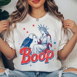 Op de afbeelding: Een wit t-shirt met korte mouwen en een afbeelding van het Vrijheidsbeeld dat de snavel van een Amerikaanse zeearend aanraakt. Het ontwerp bevat rode en blauwe sterren en het woord Boop in grote rode letters met een blauwe omlijning.