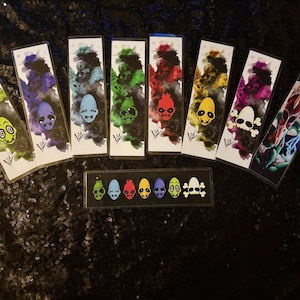 May include: A collection of colorful bookmarks featuring abstract watercolor backgrounds and stylized skull designs. Each bookmark has a unique color scheme, including green, purple, blue, red, and yellow. A smaller rectangular display case with matching skull designs is also present.