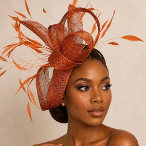Peut inclure: Un bibi orange brûlé doté d'un design en boucles de sinamay structurées et de longues plumes fines s'étendant vers l'extérieur. L'accessoire est fixé sur un serre-tête fin et inclut le texte Millinery by kukie dans le coin supérieur droit.