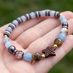 May include: A beaded bracelet with a mix of blue, brown, and gold beads. The bracelet features a dark brown star-shaped charm. The beads are of varying sizes and textures, strung on an elastic cord, creating a unique accessory.