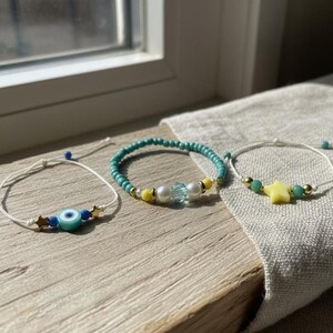 Puede incluir: Tres pulseras de cuentas sobre una superficie de madera. Una pulsera tiene un dije de ojo turco, otra tiene cuentas turquesas y la tercera tiene una estrella amarilla. Las pulseras están cerca de una ventana.