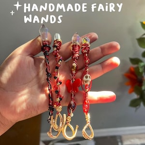 May include: Four handmade fairy wands with red and gold wire wrapped around them. Each wand has a clear crystal at the top and different charms, including a skull, a butterfly, and beads.