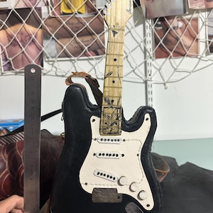 May include: A black and white leather guitar with a light brown neck. The guitar has a white pickguard and three white knobs. The headstock reads "Bensons". A ruler is next to the guitar, showing its size.