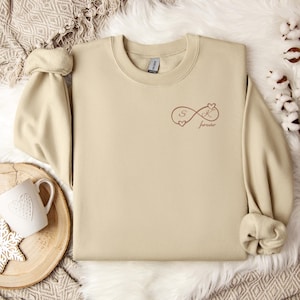 Könnte beinhalten: Beiges Sweatshirt mit hochgekrempelten Ärmeln. Das Design zeigt ein Unendlichkeitssymbol mit den Buchstaben "S" und "K" und dem Wort "forever" in Braun. Das Sweatshirt ist aus weichem Material.