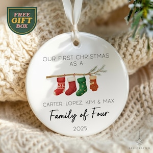 May include: White ceramic Christmas ornament with the text "OUR FIRST CHRISTMAS AS A" and "Family of Four". It features four hanging stockings in red and green. The names "CARTER, LOPEZ, KIM & MAX" and the year "2025" are also included.