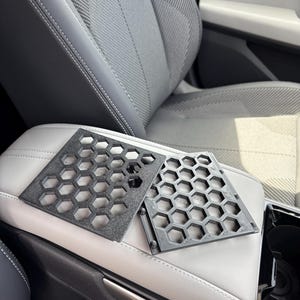 以下が含まれることがあります： ハニカム模様の2つの黒い幾何学的な車のアクセサリーが、明るい灰色の車のシートアームレストに表示されています。 アクセサリーは硬い素材でできており、正方形です。
