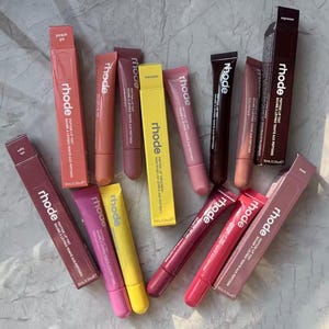 May include: An assortment of Rhode Peptide Lip Tints in shades like peach, pink, and espresso. The lip tints are presented in tubes and boxes, with the brand name visible. The products are arranged on a light-coloured surface.