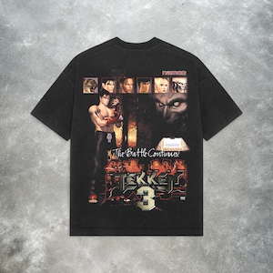 May include: Black t-shirt featuring a graphic from the Tekken 3 video game. The design includes character portraits, the game's logo, and the phrase "The Battle Continues!". The shirt has a vintage, worn look.