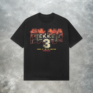 May include: Black t-shirt featuring the Tekken 3 logo in red, orange, and gray. The text "PUSH P1 OR P2 BUTTON" and "namco" are printed below the logo. The shirt has a classic crew neck and short sleeves.