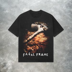 May include: Black t-shirt with a graphic of a person in a white suit lying down, and the words "FATAL FRAME" in white block letters. The image has a vintage, distressed appearance.