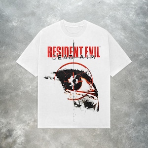 Puede incluir: Camiseta blanca con el texto "RESIDENT EVIL DEAD AIM" en rojo. El diseño presenta un ojo estilizado con una mira y detalles en negro y rojo. La camiseta tiene cuello redondo y mangas cortas.