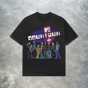 Puede incluir: Camiseta negra con un gráfico del logo de MTV y la palabra "Downtown" en blanco, sobre un fondo morado y azul. Debajo del texto hay una ilustración de dibujos animados de un grupo de personas con varios atuendos.