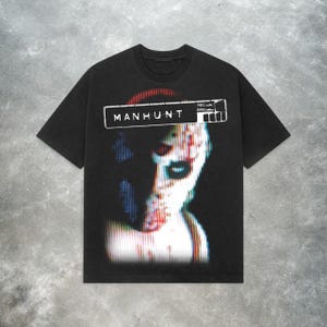 Peut inclure: T-shirt noir avec un imprim&eacute; graphique d'un visage humain avec un effet de distorsion pixelis&eacute;e. Le texte "MANHUNT" est imprim&eacute; au-dessus de l'image.