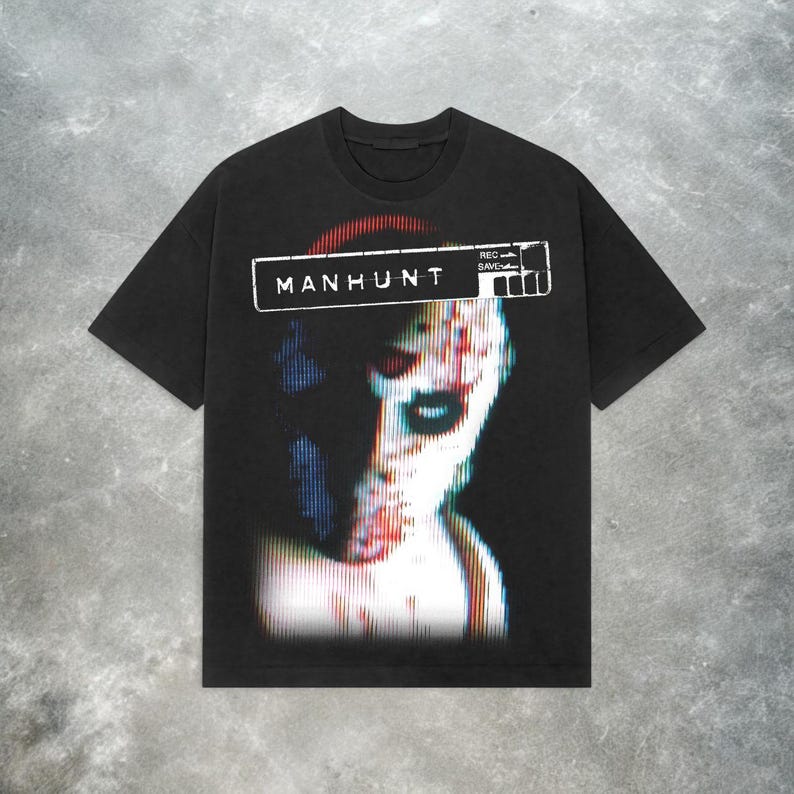 Peut inclure: T-shirt noir avec un imprim&eacute; graphique d'un visage humain avec un effet de distorsion pixelis&eacute;e. Le texte "MANHUNT" est imprim&eacute; au-dessus de l'image.