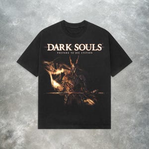 Puede incluir: Camiseta negra con un estampado gráfico de un caballero sosteniendo una espada y llamas. El texto "DARK SOULS PREPARE TO DIE EDITION" está impreso sobre la imagen.