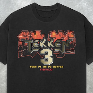 May include: Black t-shirt featuring the Tekken 3 logo in red, gold, and silver. The shirt includes the text "PUSH P1 OR P2 BUTTON" and the Namco logo. A vintage-style graphic tee.