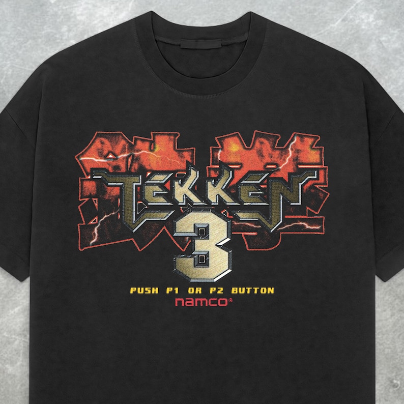May include: Black t-shirt featuring the Tekken 3 logo in red, gold, and silver. The shirt includes the text "PUSH P1 OR P2 BUTTON" and the Namco logo. A vintage-style graphic tee.