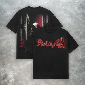 Peut inclure: Deux t-shirts noirs. L'un présente un graphisme rouge et blanc d'un personnage avec une arme à feu et le mot "Devil" en rouge. L'autre chemise porte les mots "Devil May Cry" en rouge et une silhouette de femme.