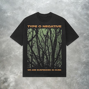 Puede incluir: Camiseta negra con el texto naranja "TYPE O NEGATIVE" sobre un gráfico de árbol verde y negro. El texto "WE ARE SUSPENDED IN DUSK" está debajo, también en naranja. La camiseta tiene cuello redondo y mangas cortas.