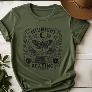 May include: An olive green t-shirt featuring a black graphic print of three moths, a stack of books, two mugs, and floral vines. The text reads MIDNIGHT READING SOCIETY with a crescent moon symbol above the moths. A brown hat is visible at the top.