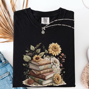 Vintage-style t-shirt featuring a stacked pile of antique books surrounded by detailed sunflowers and botanical floral accents. Perfect for book lovers and readers who enjoy a cozy, literary aesthetic. The design is printed on a soft neutral tee, ideal for fall fashion or as a thoughtful book club gift.