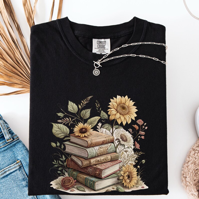 Vintage-style t-shirt featuring a stacked pile of antique books surrounded by detailed sunflowers and botanical floral accents. Perfect for book lovers and readers who enjoy a cozy, literary aesthetic. The design is printed on a soft neutral tee, ideal for fall fashion or as a thoughtful book club gift.