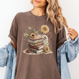Vintage-style t-shirt featuring a stacked pile of antique books surrounded by detailed sunflowers and botanical floral accents. Perfect for book lovers and readers who enjoy a cozy, literary aesthetic. The design is printed on a soft neutral tee, ideal for fall fashion or as a thoughtful book club gift.