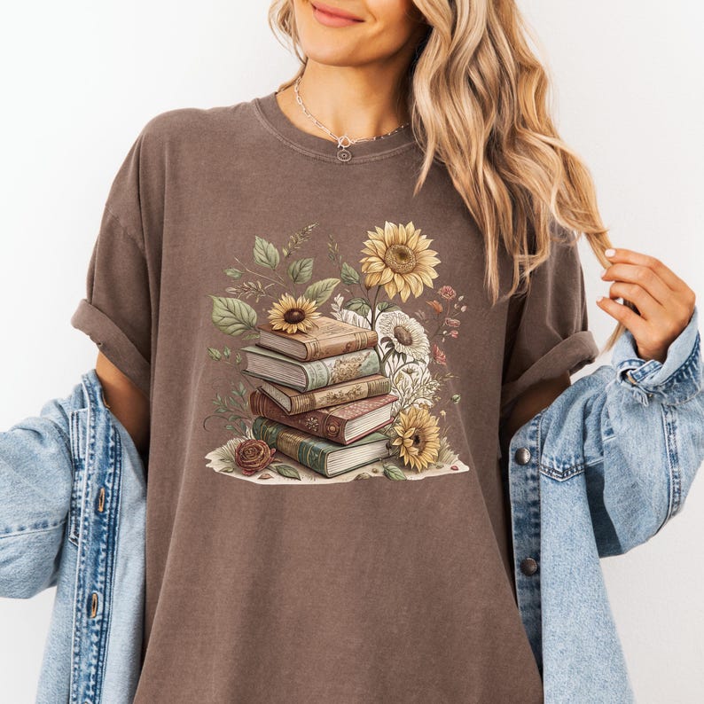 Vintage-style t-shirt featuring a stacked pile of antique books surrounded by detailed sunflowers and botanical floral accents. Perfect for book lovers and readers who enjoy a cozy, literary aesthetic. The design is printed on a soft neutral tee, ideal for fall fashion or as a thoughtful book club gift.