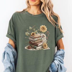 Vintage-style t-shirt featuring a stacked pile of antique books surrounded by detailed sunflowers and botanical floral accents. Perfect for book lovers and readers who enjoy a cozy, literary aesthetic. The design is printed on a soft neutral tee, ideal for fall fashion or as a thoughtful book club gift.