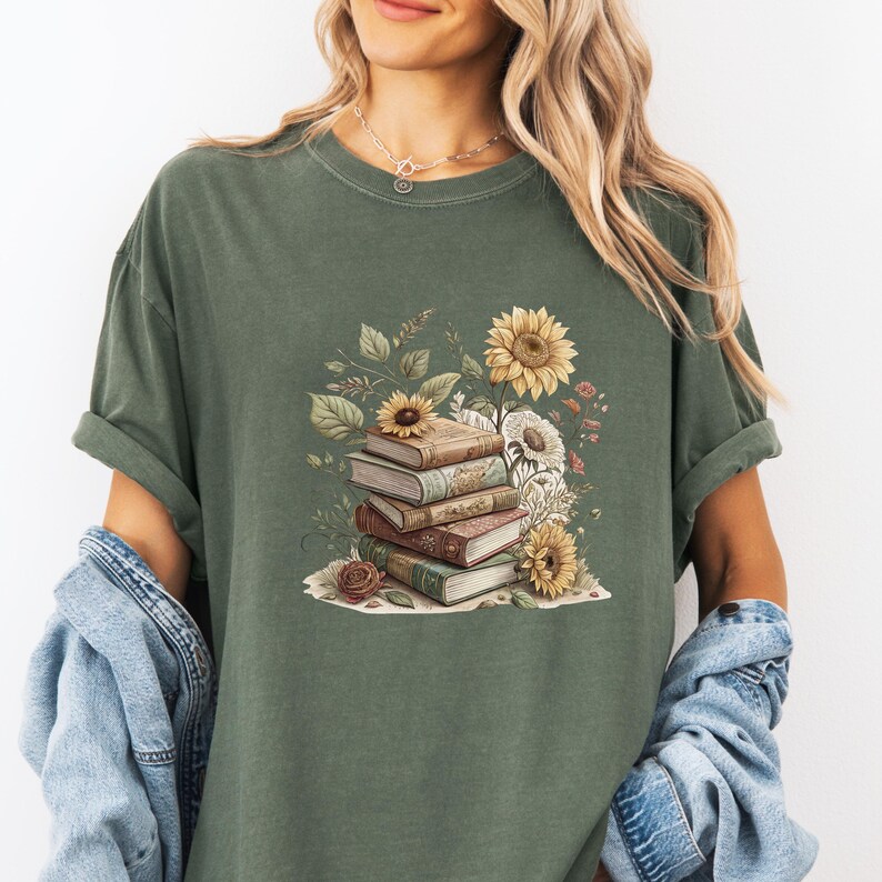 Vintage-style t-shirt featuring a stacked pile of antique books surrounded by detailed sunflowers and botanical floral accents. Perfect for book lovers and readers who enjoy a cozy, literary aesthetic. The design is printed on a soft neutral tee, ideal for fall fashion or as a thoughtful book club gift.