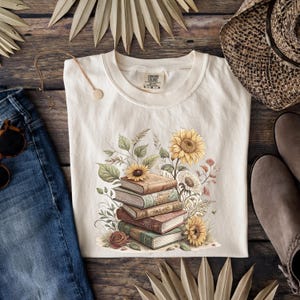 Vintage-style t-shirt featuring a stacked pile of antique books surrounded by detailed sunflowers and botanical floral accents. Perfect for book lovers and readers who enjoy a cozy, literary aesthetic. The design is printed on a soft neutral tee, ideal for fall fashion or as a thoughtful book club gift.