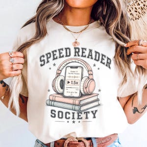 Peut inclure: T-shirt crème avec un motif graphique. Le motif comprend les mots "SPEED READER SOCIETY", un casque audio, un smartphone affichant un podcast et deux livres. Le design est dans les tons beige, rose et gris.