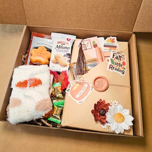 May include: A gift box filled with fall-themed items. Includes a book wrapped in brown paper, a pair of fuzzy socks, snacks, and decorative accents like stickers and flowers. The box is open, revealing the contents.