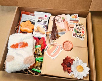 May include: A gift box filled with fall-themed items. Includes a book wrapped in brown paper, a pair of fuzzy socks, snacks, and decorative accents like stickers and flowers. The box is open, revealing the contents.