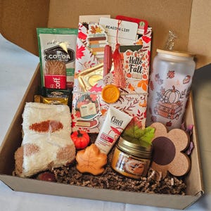 May include: A gift box filled with fall-themed items. Includes a cocoa packet, chocolate bars, a reading list, a candle, a tumbler with a pumpkin design, a pair of socks, a leaf-shaped soap, and a tube of lotion. The box is lined with brown paper.
