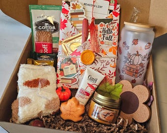 May include: A gift box filled with fall-themed items. Includes a cocoa packet, chocolate bars, a reading list, a candle, a tumbler with a pumpkin design, a pair of socks, a leaf-shaped soap, and a tube of lotion. The box is lined with brown paper.