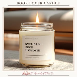 May include: A lit soy wax candle in a glass jar. The candle has a white label with the text "SMELLS LIKE BOOK HANGOVER". The candle is 19 oz. The brand name is Bookmarked Bliss Co.