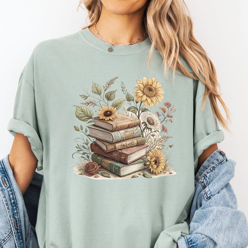 Vintage-style t-shirt featuring a stacked pile of antique books surrounded by detailed sunflowers and botanical floral accents. Perfect for book lovers and readers who enjoy a cozy, literary aesthetic. The design is printed on a soft neutral tee, ideal for fall fashion or as a thoughtful book club gift.
