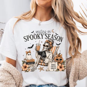 May include: A white t-shirt featuring a graphic print with the text "waiting on SPOOKY SEASON". The design shows a skeleton wearing a headband and sunglasses, reading a book titled "ONE MORE CHAPTER", with a ghost, pumpkin, and tote bag.