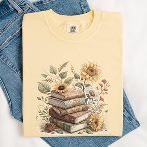 Vintage-style t-shirt featuring a stacked pile of antique books surrounded by detailed sunflowers and botanical floral accents. Perfect for book lovers and readers who enjoy a cozy, literary aesthetic. The design is printed on a soft neutral tee, ideal for fall fashion or as a thoughtful book club gift.