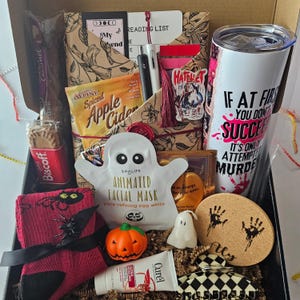 May include: A Halloween-themed gift box filled with various items. Includes a white tumbler with a quote, a ghost-shaped face mask, a pumpkin-shaped soap, a reading list, snacks, and a black and white hair clip. The box is decorated with a red and black theme.