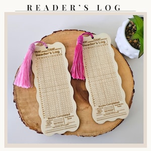 May include: Two wooden Reader's Log bookmarks with pink tassels, displayed on a wooden slice. Each bookmark has a grid for tracking books read, with the text "Reader's Log" at the top. The bookmarks are light brown with dark brown text.