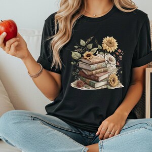 Vintage-style t-shirt featuring a stacked pile of antique books surrounded by detailed sunflowers and botanical floral accents. Perfect for book lovers and readers who enjoy a cozy, literary aesthetic. The design is printed on a soft neutral tee, ideal for fall fashion or as a thoughtful book club gift.