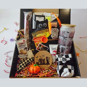 May include: A Halloween-themed gift box filled with various items. Includes a black cat-shaped container, a glass with a ghost and book design, a pumpkin, a coaster with a cat silhouette, and a package of Milano cookies. The box is decorated with orange and black filler.