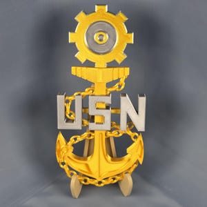 Puede incluir: Un emblema de ancla dorado con las letras "USN" en plata, conectadas por una cadena. Encima de las letras hay un diseño de engranaje dorado. El emblema se muestra sobre un pequeño soporte de madera sobre un fondo gris.