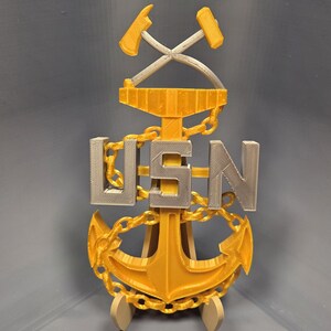 May include: A 3D-printed USN Chief Petty Officer insignia in gold and silver. The design includes crossed hammers, the letters "USN", and an anchor. The insignia is mounted on a small stand.