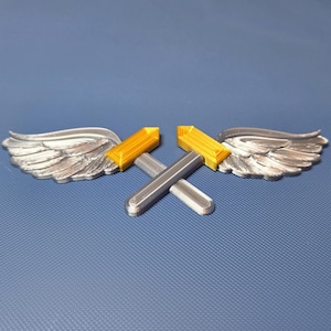 May include: Two silver-colored wings with a gold and silver hammer design. The wings are detailed with feather textures. The hammers are crossed and have a gold head. The background is a solid blue color.