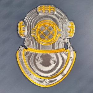 May include: A silver and gold-colored diving helmet with a circular faceplate and a curved neckpiece. The helmet features intricate details, including rivets and a grid pattern on the faceplate. The design is a 3D-printed object.