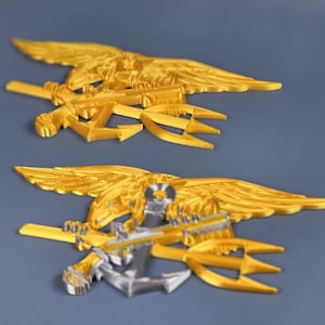 Op de afbeelding: Twee goudkleurige metalen emblemen, elk met een gespreide adelaar die een drietand en een horizontaal object vasthoudt. Het onderste embleem bevat een zilveren anker. De achtergrond is effen, gedempt blauw.